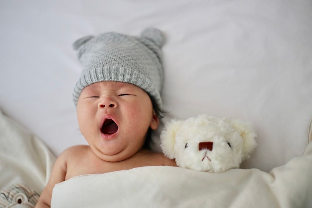 a baby wearing a grey sock hat yawning, covered in a white blanket