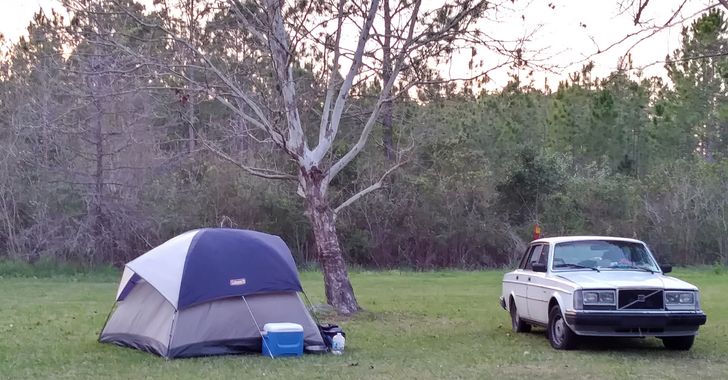 A blue tent under a tree with a cooler in front of it and a white sedan next to it
