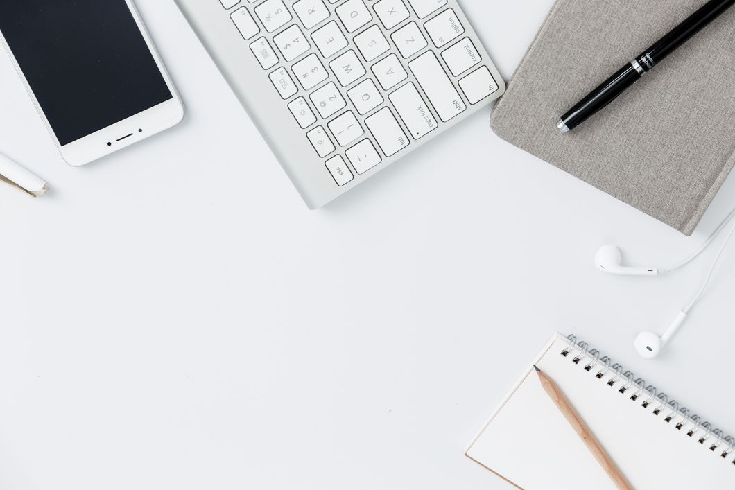 A close-up stock image of a desk and its contents. There's an iPhone, a keyboard for a Mac computer, two notebooks, a pen, a pencil and Mac headphones with cords.