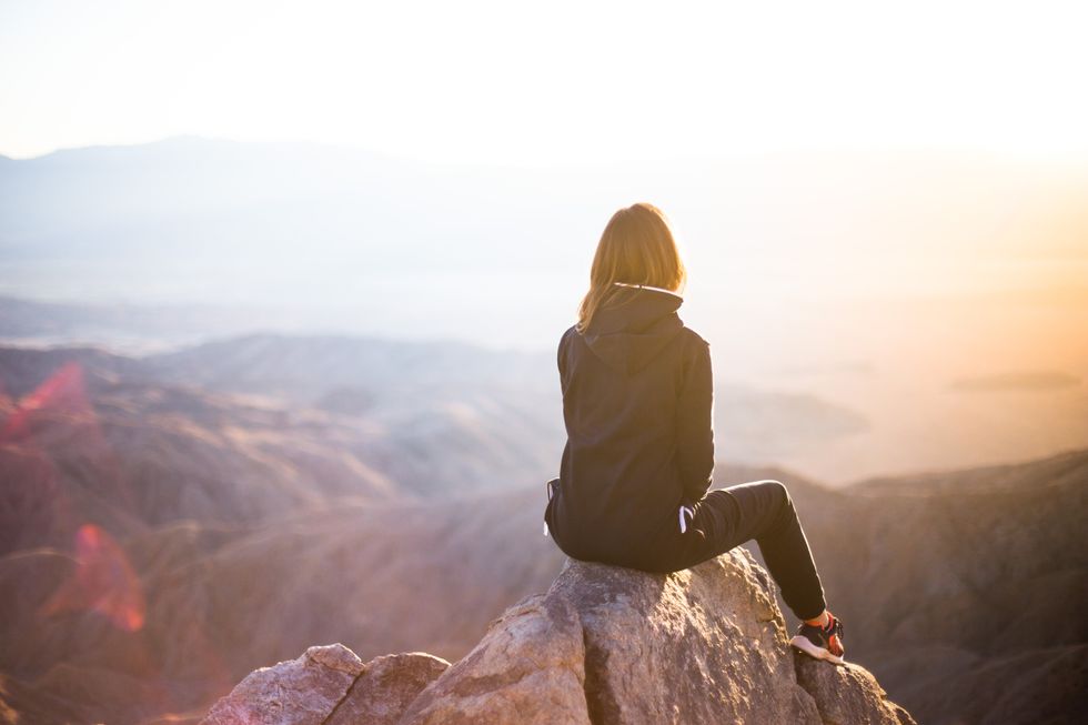 A girl is staring at a mountian view she is wearing a black jacket black pants and what appears to be orange and black shoes