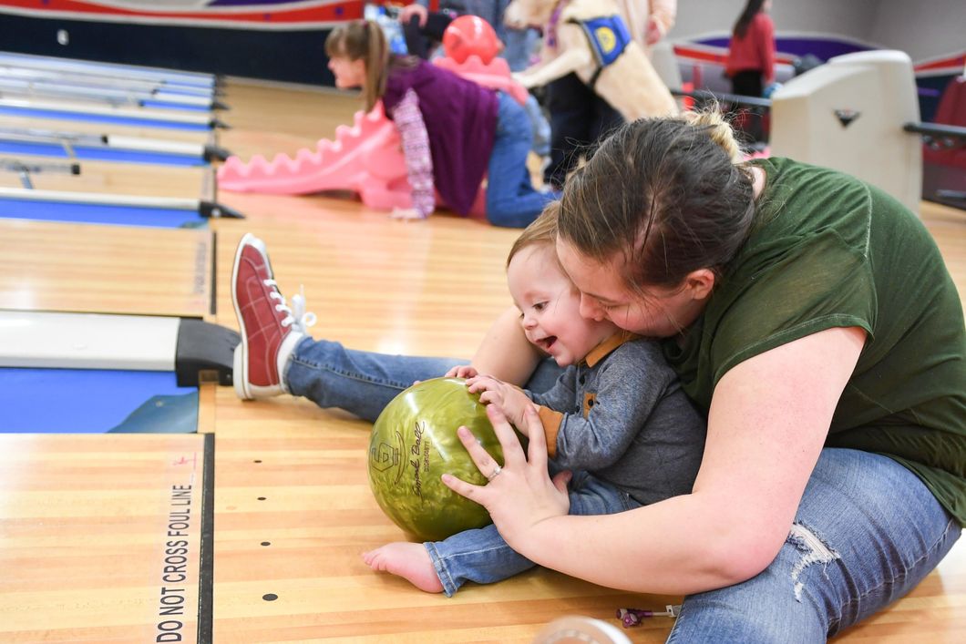 A lady and a child are sitting on a floor in a bowling alley they are holding a green bowling ball
