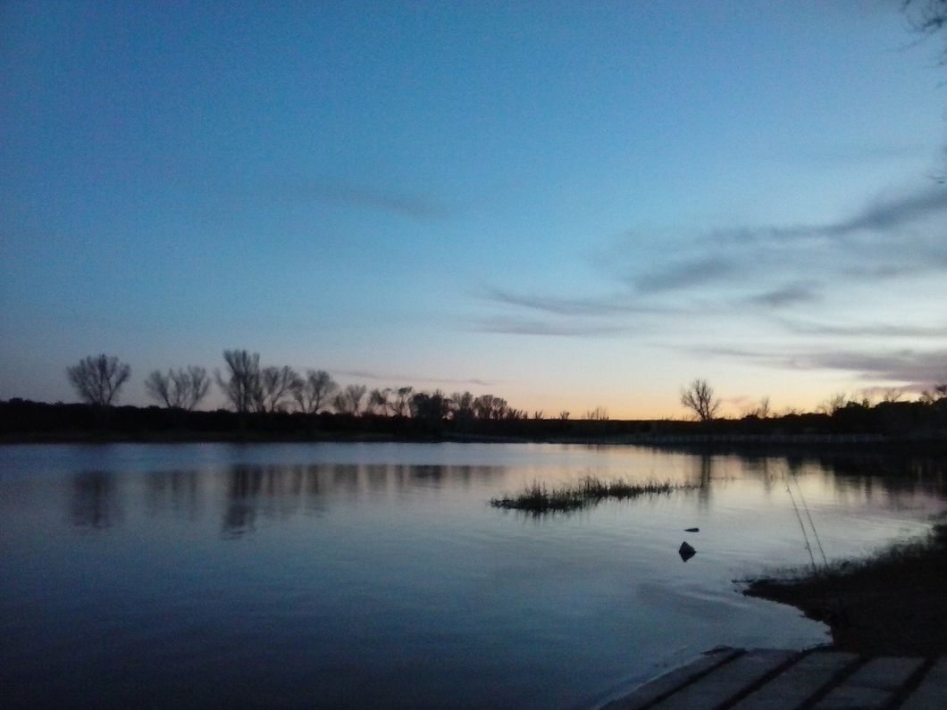 A lake at dusk, the sky and the lake are almost the same shade, light is barely remaining in the sky as the sun sets. Two fishing poles are resting in an upright position at the right 