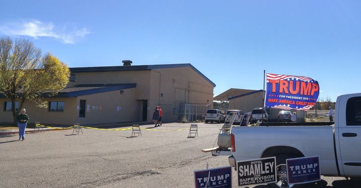 A large brown building on the left and a white pickup on the right with a Trump election sign surrounded by local election signs