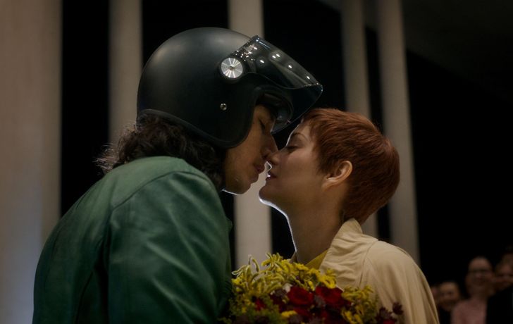 A movie still from "Annette." It's a tight framed movie still of the actors' side profiles as they lean in for a kiss. Adam Driver (left) wears a green leather jacket and motorcycle helmet. Marion Cotillard (right) wears a bright yellow dress with yellow flowers in hand.