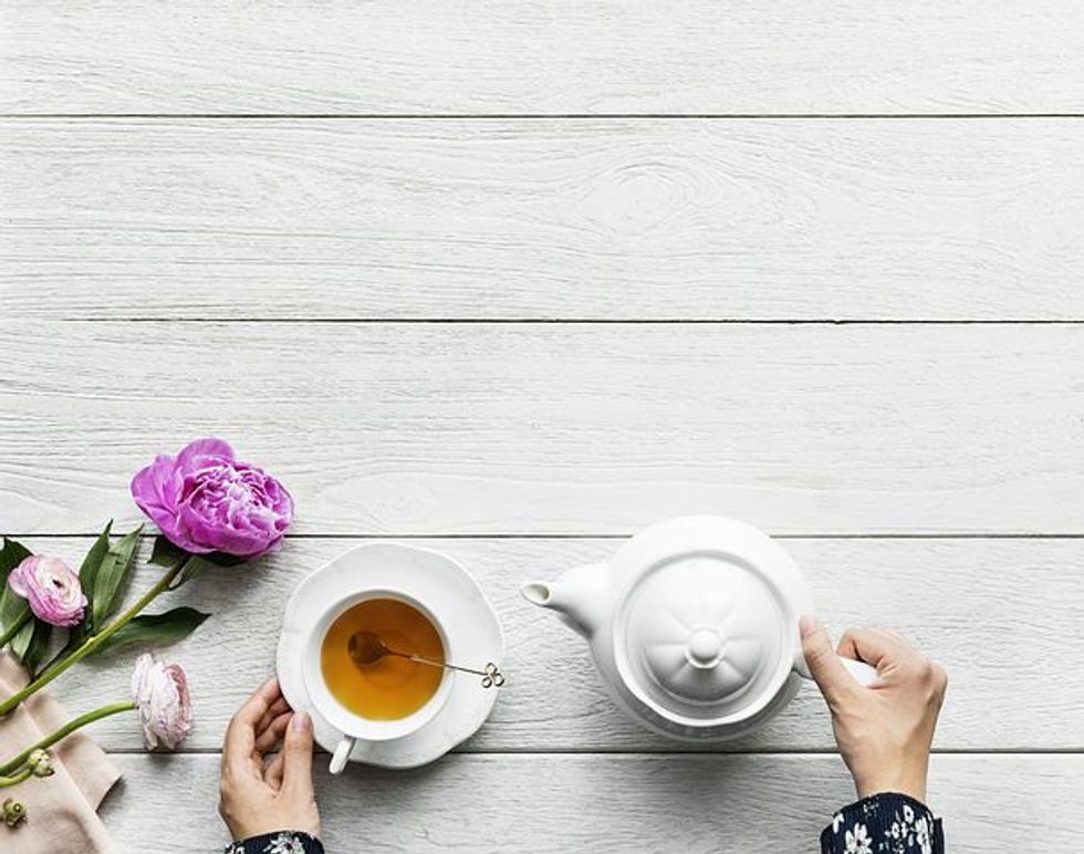 A person pouring tea from a white teapot into a cup on a wooden table with pink peonies.