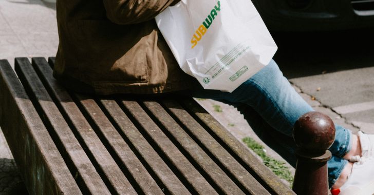 a person sitting on a bench with a subway bag of food
