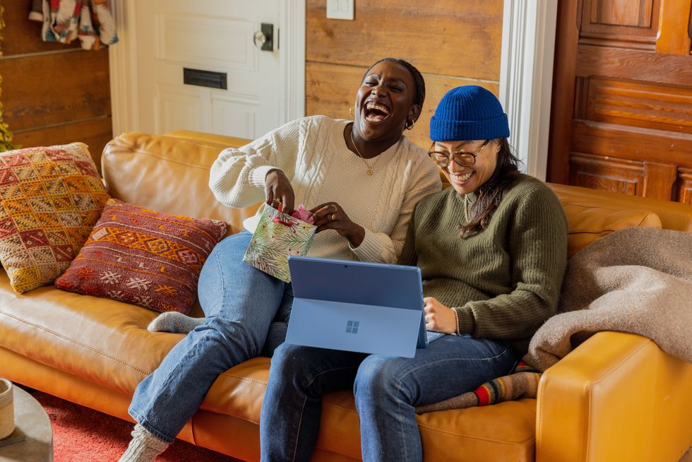 a person sitting on a couch with a laptop while another one is laughing and opening a bag right next to them