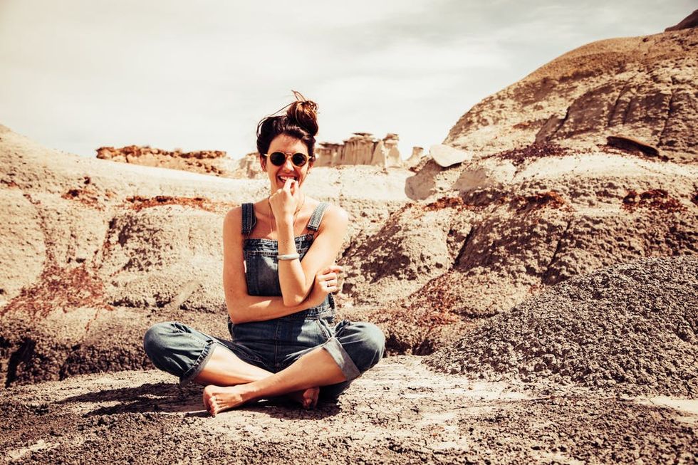 A person sitting on rocky terrain, wearing sunglasses and overalls, smiling playfully.