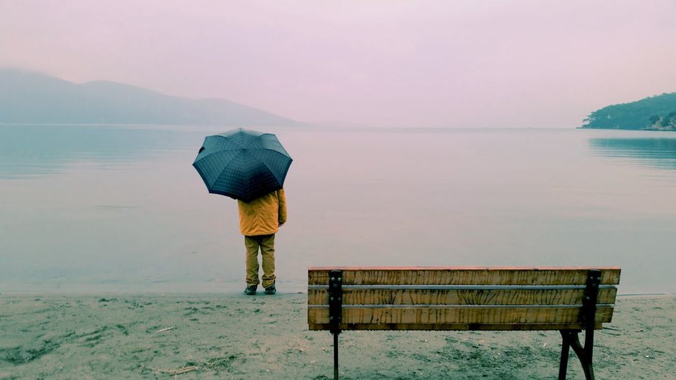 A person with an umbrella stands by a calm lake with distant hills, facing away. An empty wooden bench is in the foreground.