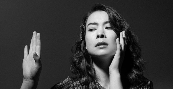A photo of Mitski looking to the left with her hand up.