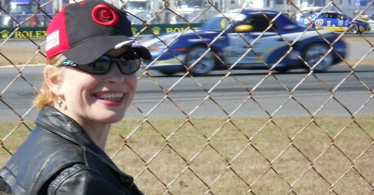 A red-headed woman with a black Target hat on smiling standing in front of a fence with a blue race car behind the fence