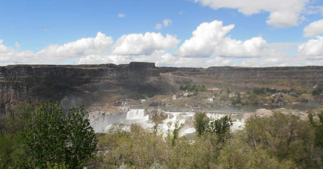 A rocky canyon with a distant but huge waterfall in the center with trees and greenery in the foreground