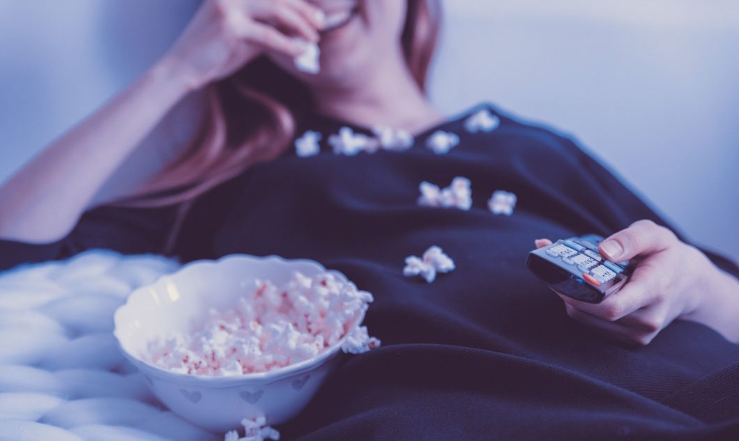 A stock image of a woman wrapped in a blanket, eating popcorn with her right hand and using a TV remote in her left hand. She's smiling, but the photo cuts off her nose and above.