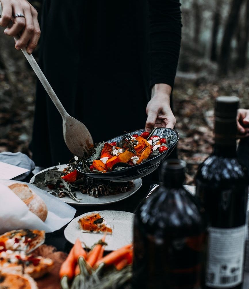 a table covering in orange and black festive foods and wine bottles with a person in all black filling their plate.