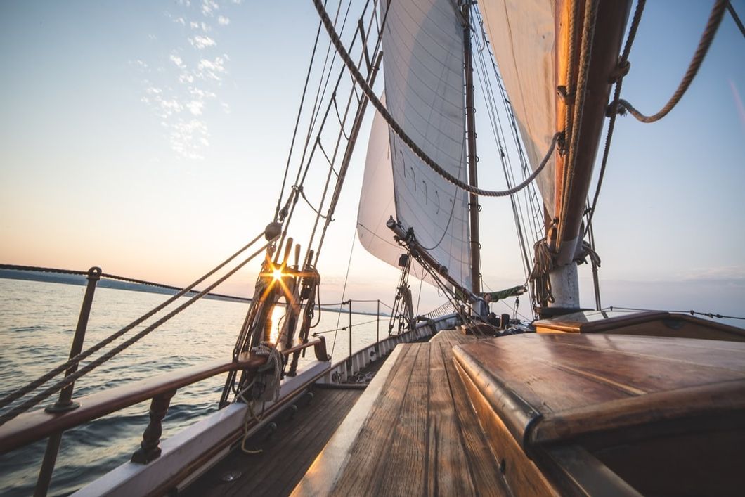  a view of the front of a wooden sailboat from the point of view of someone sitting to the left of the cockpit during sunrise.