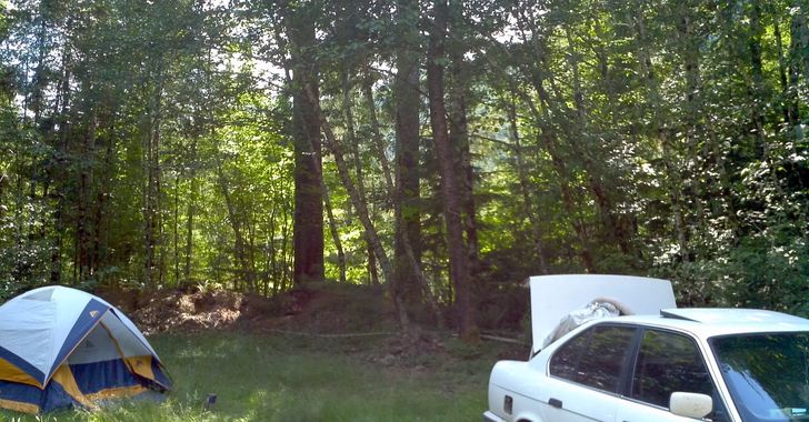A white, blue and yellow tent on a grassy area in front of woods with a white sedan with a trunk open in the foreground