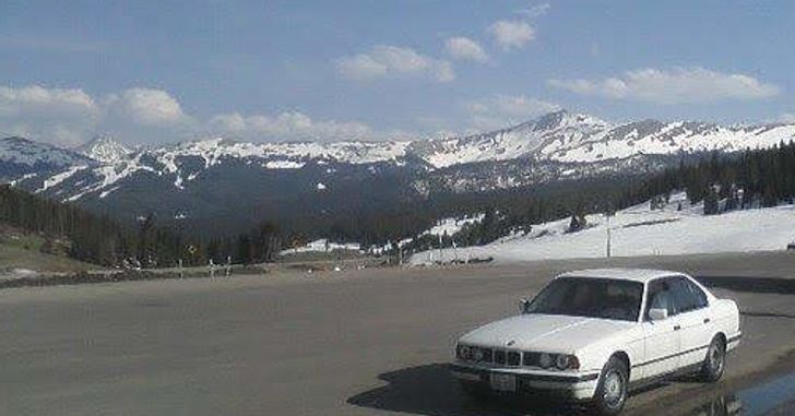 A white BMW sedan on a rest stop concrete in the snow in the Rocky Mountains with snow-covered peaks behind a few trees