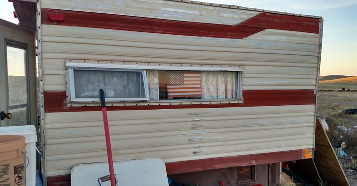 a white with red stripes homemade pickup camper sitting on the ground with the back door open.