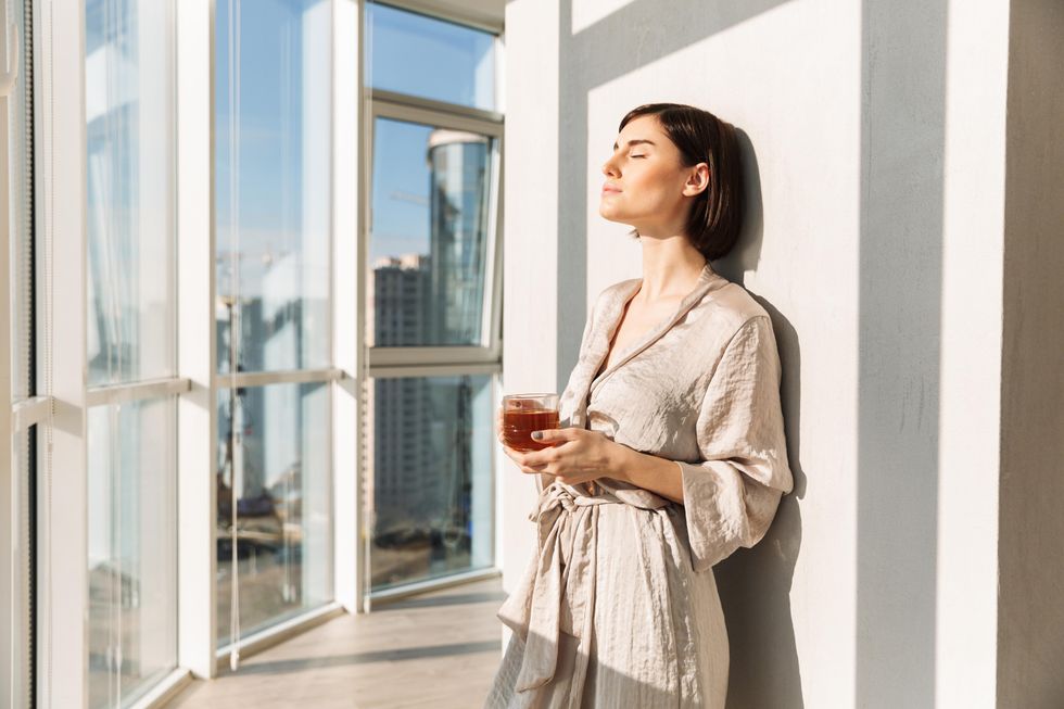 A woman standing, resting in the sunlight holding a cup of red liquid