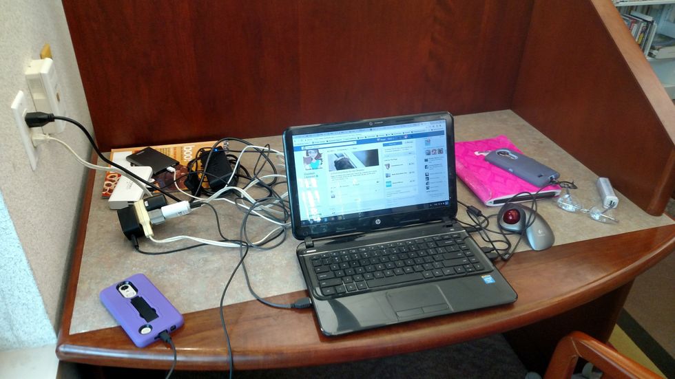 A wooden cubical with a laptop and many cords attached to an outlet with phones and batteries plugged in