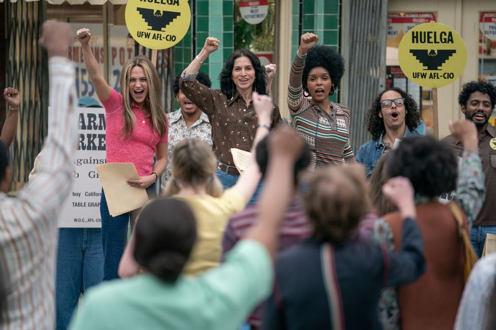​Alicia Vikander, Monica Sanchez and Janelle Monáe in THE GLORIAS. All three ladies are standing in the background with their fists in the air. The out of focus crowd is also lifting up their hands. 
