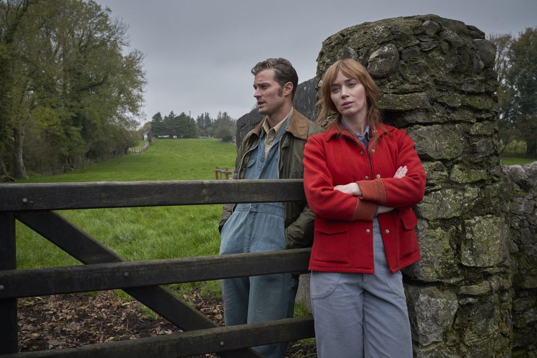 amie Dornan (Left) stands beside Emily Blunt (Right) with a wooden gate separating the two. Both face the camera, but look past it at the Irish, green landscape. The sky is dreary and grey. He wears farming overalls and a collared brown shirt with a large jacket. She wears a red jacket and similar farm overalls.