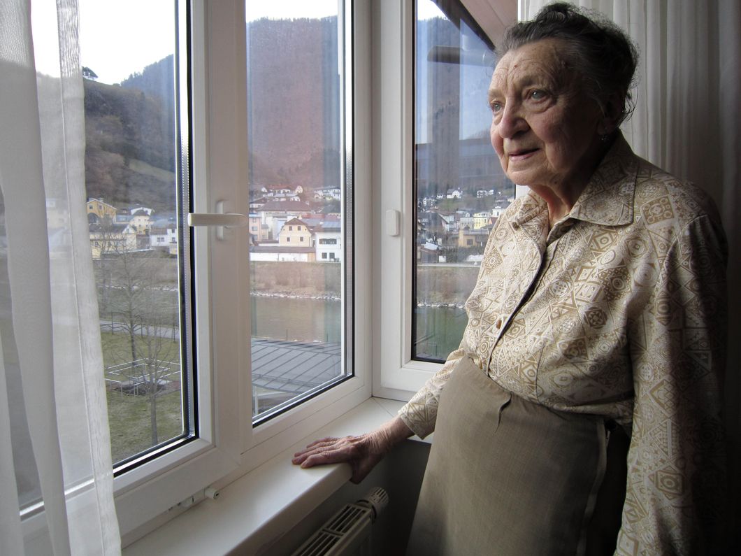 An elderly woman stands near a window with curtains open, overlooking a town in Germany. She is one of many individuals who offer first-hand accounts of living in Germany during Adolf Hitler's Third Reich.
