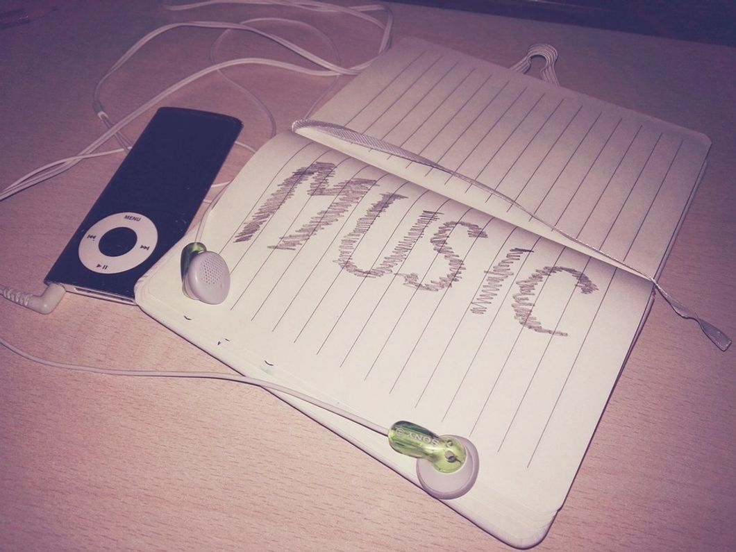 An open notebook with the word MUSIC scribbled in large letters, and an old iPod with a pair of earbuds sitting to the side.