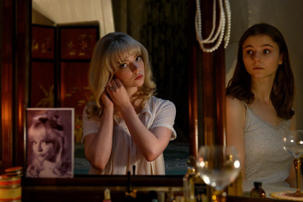Anya Taylor-Joy as Sandie puts on earrings and stares at a dresser's mirror while Thomasin McKenzie as Eloise looks on in an adjacent reflection. A photo of Sandie sits on the mirror in this still image from Focus Features' "Last Night in Soho."