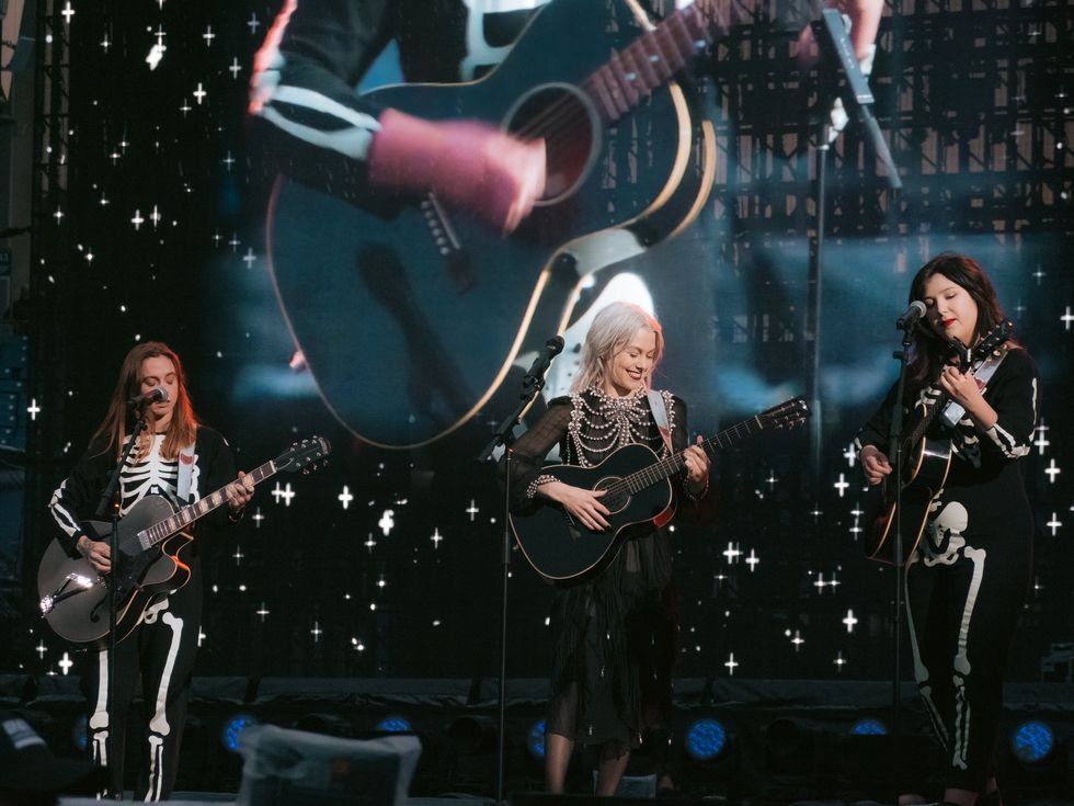 boygenius in skeleton outfits perform with guitars on a stage with starry backdrop.