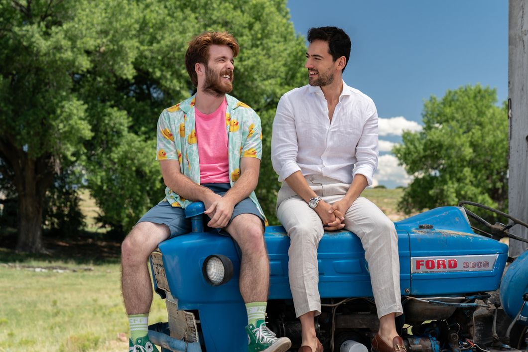Connor Del Rio (left) as "Asher" and Luis Gerardo Mendez (right) as "Renato" in "Half Brothers." Connor is wearing a button down short sleeve shirt with rubber ducks on them and a pink shirt underneath it. Blue shorts and green socks and converse. Luis is wearing a dress shirt, slacks and moccasins. They're looking at each other, laughing and sitting on a small farming tractor. In the background, it's green and bright. It looks like a farm.