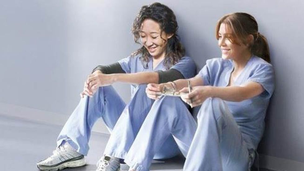 Cristina Yang and Meredith Grey in scrubs sit against a wall, smiling and enjoying a break.