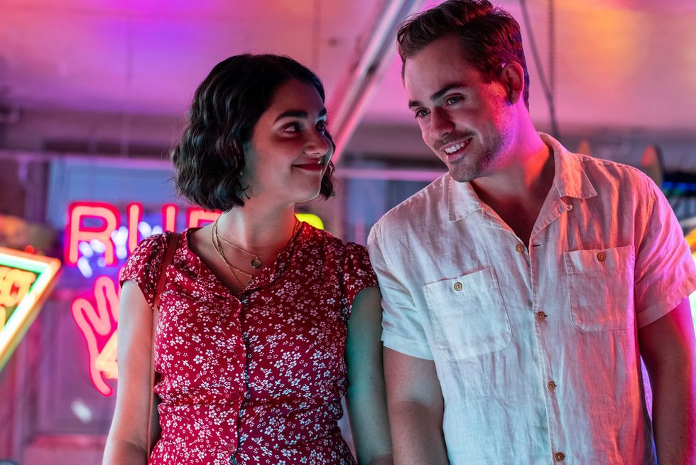 Dacre Montgomery and Geraldine Viswanathan are pictured in a pink-saturated neon sign shop. Geraldine is smiling at Dacre while he looks on at a sign. She wears a red dress with flowers, and he wears a white short-sleeved button down shirt.