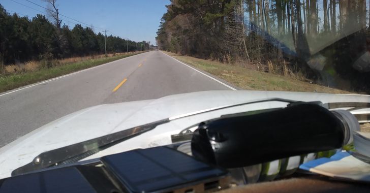 Dashboard of a white car with a small solar panel and an electrical tape covered plastic bottle and a two lane road ahead of it