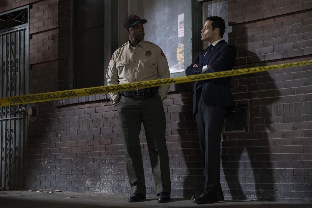 Denzel Washington (left) and Rami Malek (right) stand behind a caution tape line against a brick wall in John Lee Hancock's crime thriller "The Little Things." The ambiance of the photo is dark, bleak and mysterious due to low lighting and exposure.