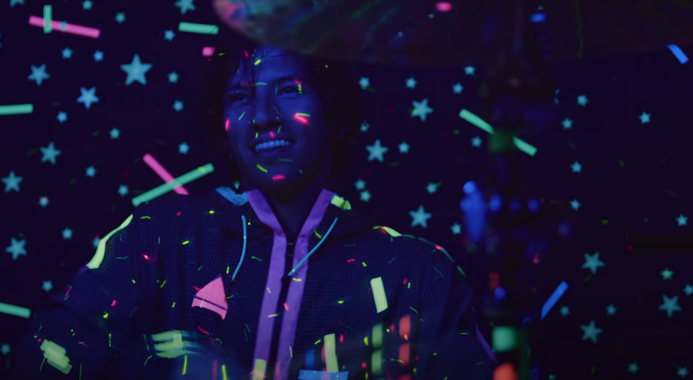 Drummer Josh Dun from Twenty One Pilots smiles while performing in the "Level of Concern" music video. He is sitting in the dark as neon lights dance across his body and stars glow on the wall behind him.