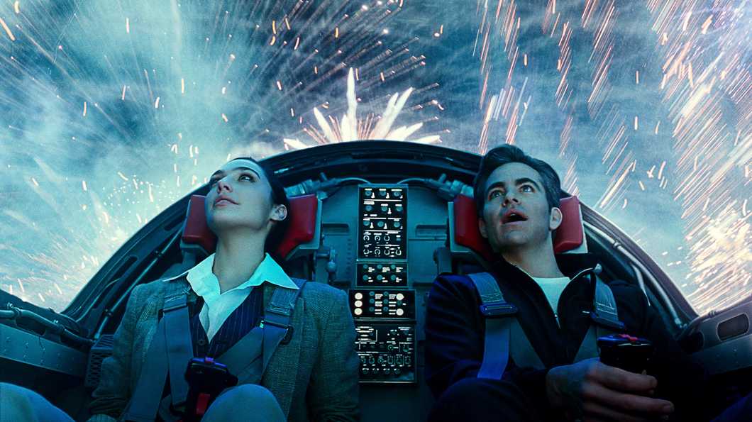 Gal Gadot as Diana Prince (left) and Chris Pine as Steve Trevor (right) take a ride in an airplane on Fourth of July. The photo is of the couple angled upwards so viewers can see fireworks light up the sky above them as they sit in the cockpit.