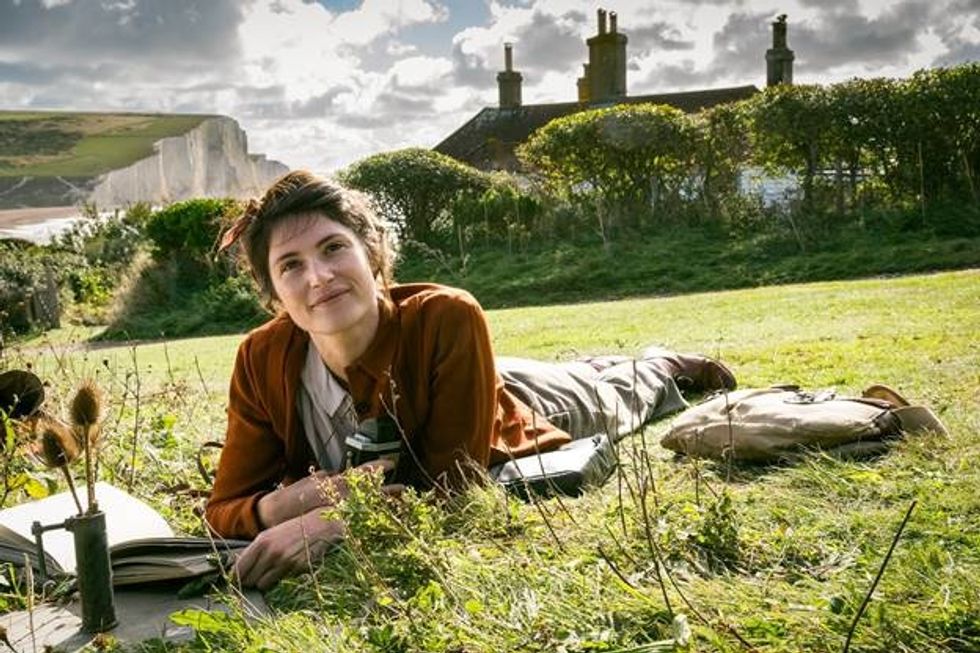 ​ Gemma Arterton plays Alice, a reclusive writer in Southern England, in British World War II drama "Summerland." She is lying belly down on the grass atop a hill with a book in her hand. She is smiling as her gaze is looking up.