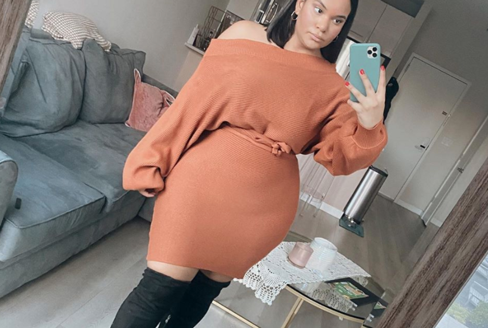 Girl wearing orange sweater dress and black over-the-knee boots taking mirror selfie.