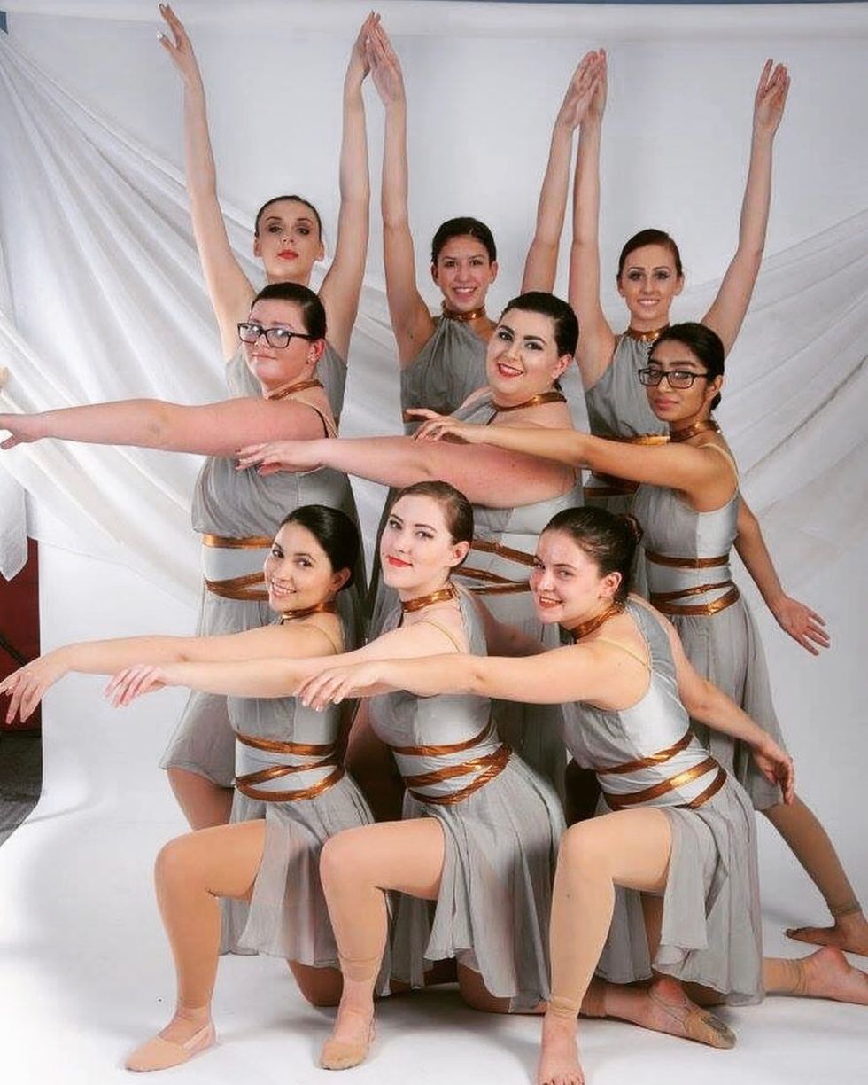 Group of dancers in gray costumes with gold ribbons, striking a synchronized pose.