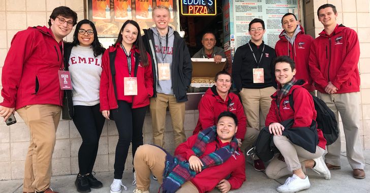 Group of ten people in red jackets posing outside a pizza shop.