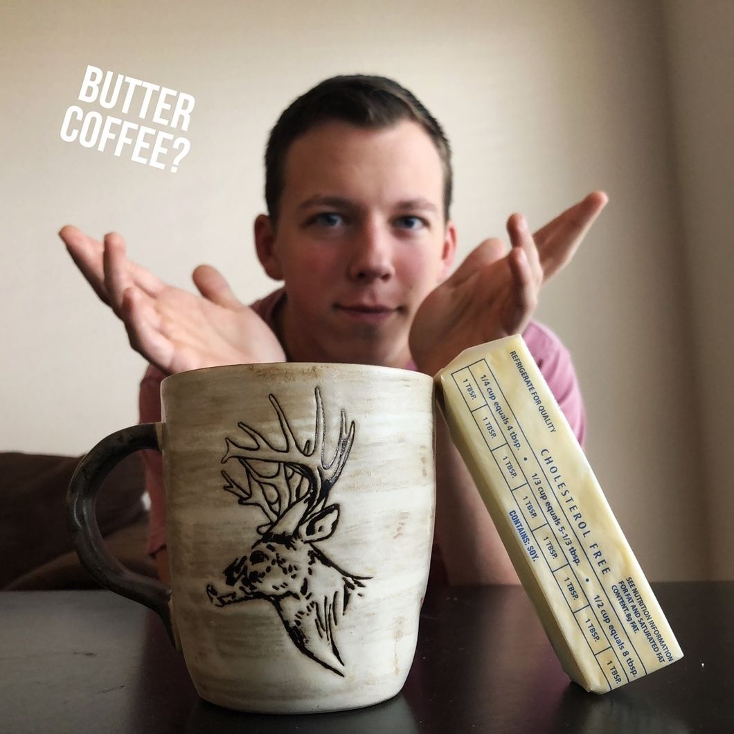 Guy looking at the camera up close, with a cup of coffee and a stick of butter in front of him. He is holding his hand out, with a questioning demeanor on face, with the caption "butter coffee" positioned to his left as a graphic