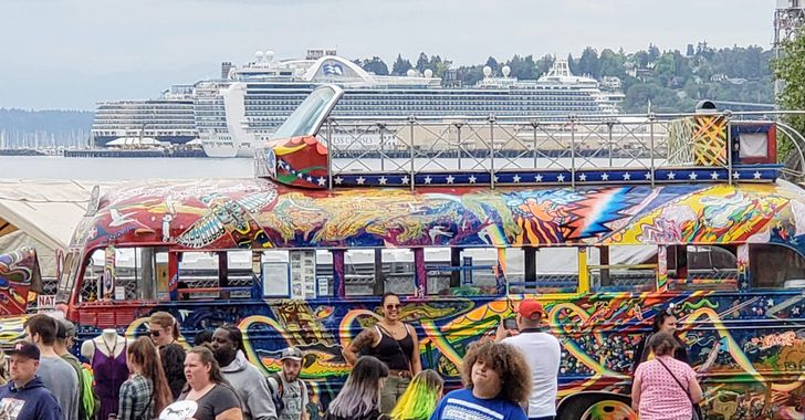 Image: A multicolored painted bus behind a crowd of people milling about all in front of Elliott Bay with 2 cruise ships docked
