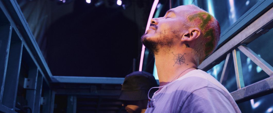 J Balving has his eyes closed and his head lifted up to the sky. The photo is taken from his left side, so his compass tattoo is shown on the left side of his neck. He also has hair paint on his buzz cut head, and it says "Amor" in lime green letters highlighted by red tints.