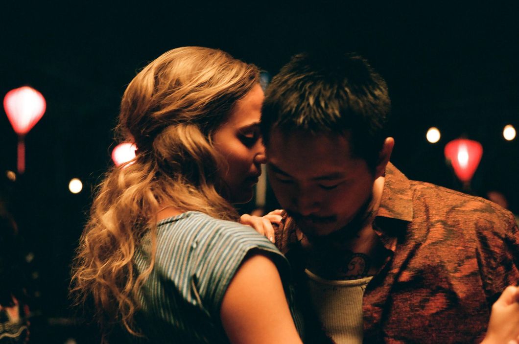 ​Kathy (Alicia Vikander) and Antonio (Justin Chon) dance during a dinner party scene in the film "Blue Bayou." Kathy is wearing a green-white striped dress while Antonio wears an open red shirt with a white tank underneath. The photo is a tight, intimate shot of the couple dancing in each other's arms. 