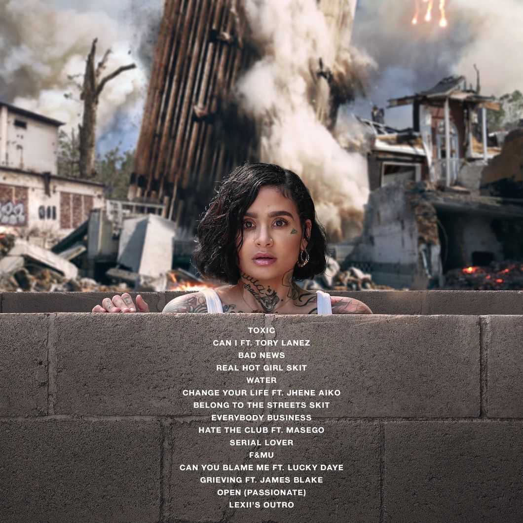 Kehlani looks over a brick wall and stares in disbelief as a destroyed building sits behind her
