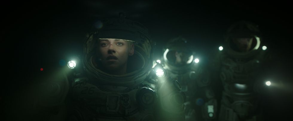 Kristen Stewart is in an underwater diver or spacesuit pictured in the foreground with her only source of light coming from her lamps on the suit. There are two other actors blurred in the background behind her. They walk on the ocean floor in darkness.