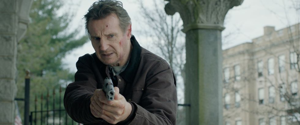 Liam Neeson as Tom Carter in "Honest Thief." He's wearing a brown jacket and about two additional layers of clothing. He's pointing a pistol toward a suspect on the floor. In the background, he's in a suburban area with iron fences and both leaf-less and full trees.