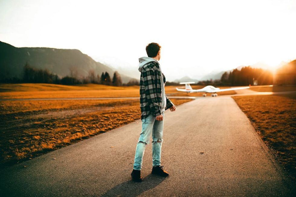 Man standing on a road at sunset with a small airplane and mountains in the background.