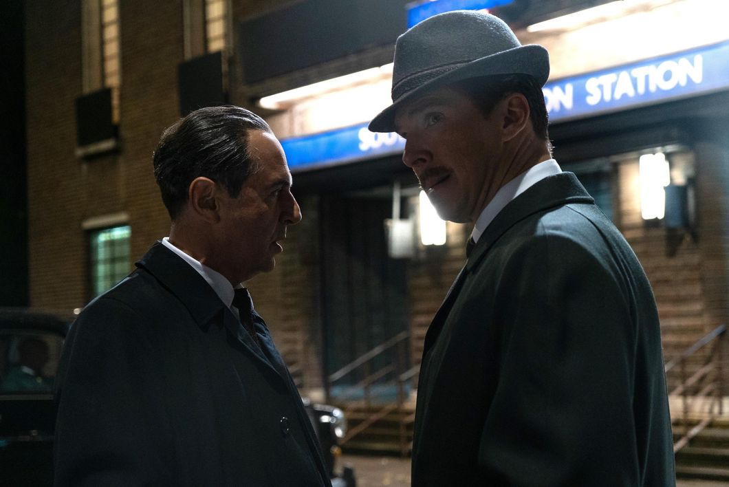 Merab Ninidze (standing left) and Benedict Cumberbatch (standing to the right, looking to the left) in a movie still photo from "The Courier." Both men are wearing dark suits. It's the early 1960's, and the men are standing outside on a dark street in front of a train station entrance.